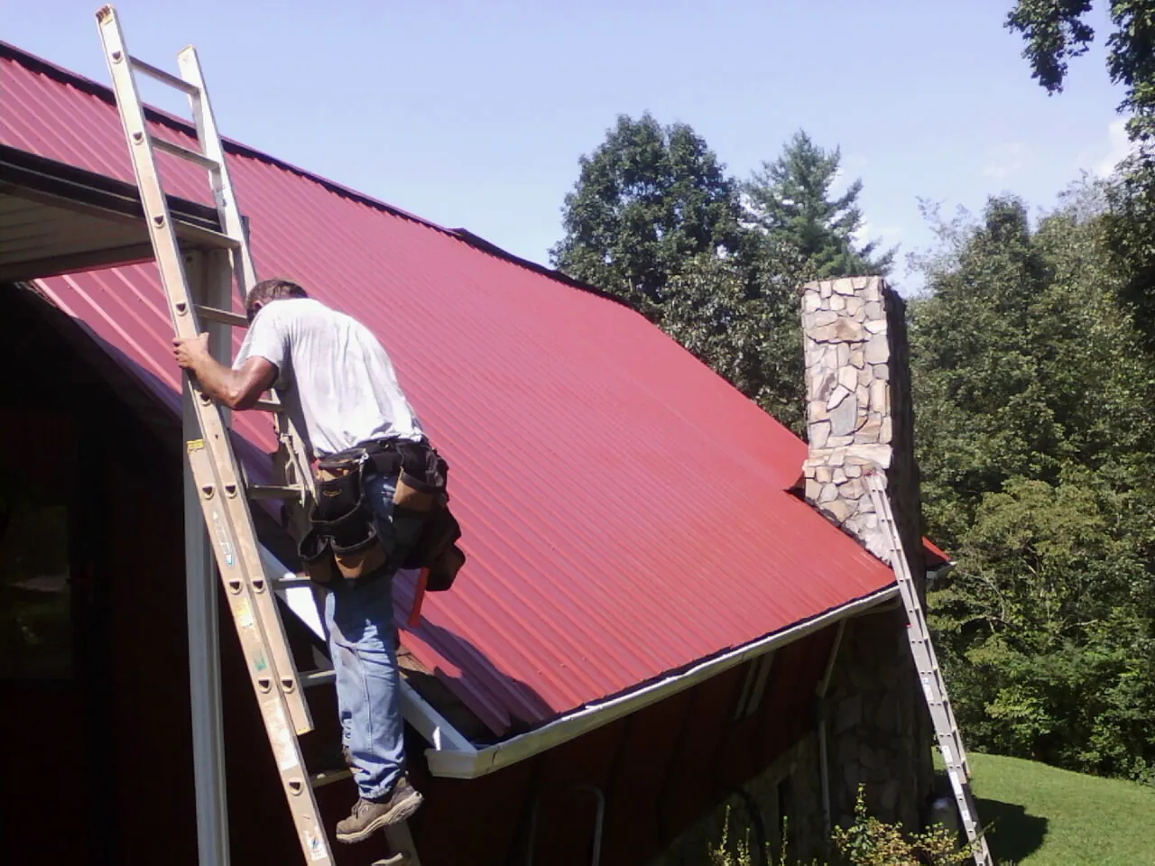 Roofer on a ladder beside a red metal roof and stone chimney — Soffit & Fascia Repair by Roofing Brothers in Narragansett